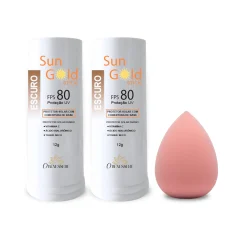 Dois bastões de protetor solar Sun Gold FPS 80 do tom Escuro, 12 g, ao lado de uma esponja de maquiagem rosa
