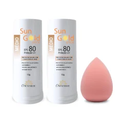 Dois bastões de protetor solar Sun Gold Stick FPS 80 tom Médio, de 12 g, ao lado de uma esponja de maquiagem cor de rosa